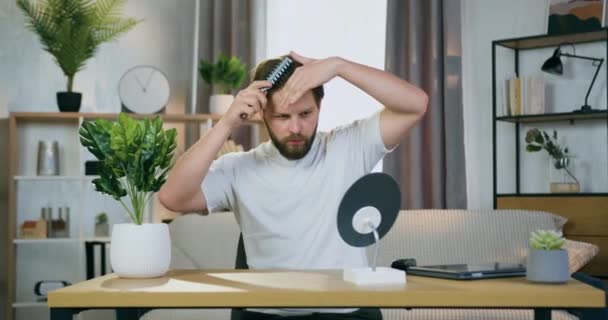 Привлекательный уверенный молодой бородатый мужчина в белой футболке расчесывает волосы, сидя за столом перед маленьким зеркалом в современной комнате — стоковое видео