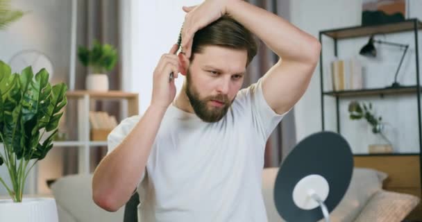 Likable concentrated young bearded guy in homewear sitting in front of small mirror in cozy room and combing his hair — Stock Video