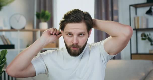 Close up of good-looking satisfied confident young bearded guy in homewear which poses at camera while making stylish hairstyle at home — Stock Video