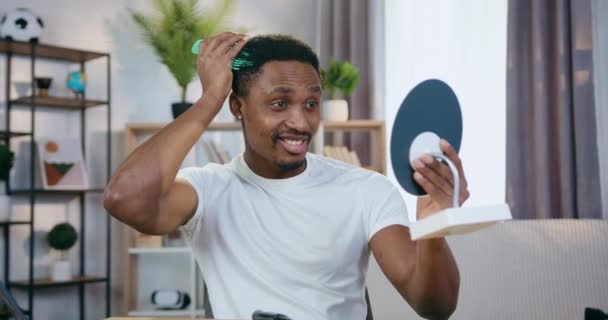 Likable joyful confident young black-skinned guy looking in small mirror and combing his hair and beard with special comb,slow motion — Stock Video