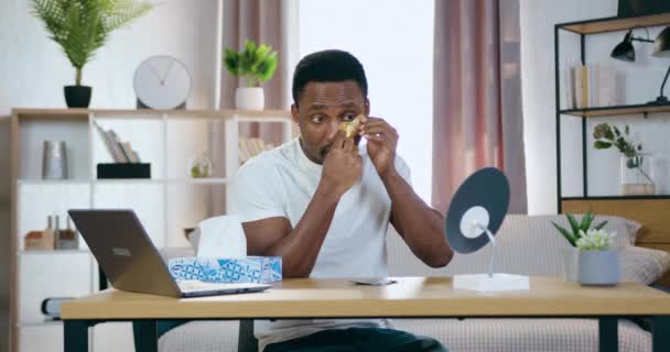 Knap positief jong Afrikaans amerikaans zitten in de voorkant van kleine spiegel thuis en zet gouden patchi onder ogen, gezichtsverzorging concept — Stockvideo