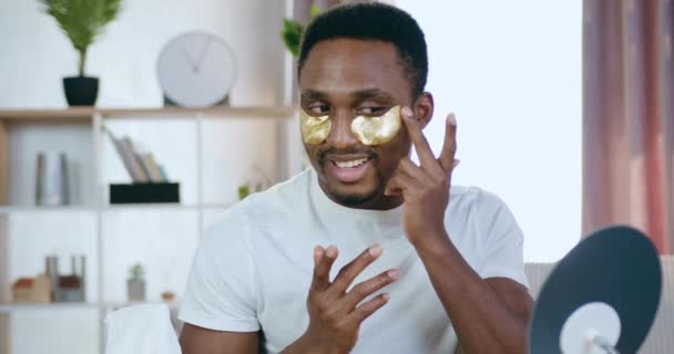 Beauty concept waar goed uitziende glimlachende jonge moderne zwart-huid man zitten voor de spiegel thuis en zet eye-patches — Stockvideo