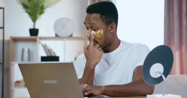 Atractivo cuidado positivo joven de piel negra chico con parches de ojos dorados sentado en la mesa en casa y trabajando en la computadora, concepto de cuidado de la piel — Vídeos de Stock