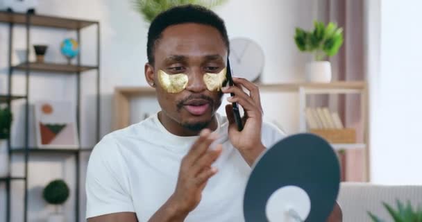 Portrait of good-looking smiling modern young black-skinned man which putting golden patches under eyes to refresh face skin and talking on mobile at home — Stock Video