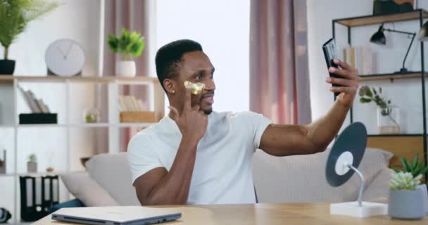 Guapo sonriente afroamericano satisfecho mirando a la cámara del teléfono y mirando a su reflejo con parches de hidrogel, concepto de cuidado facial — Vídeos de Stock