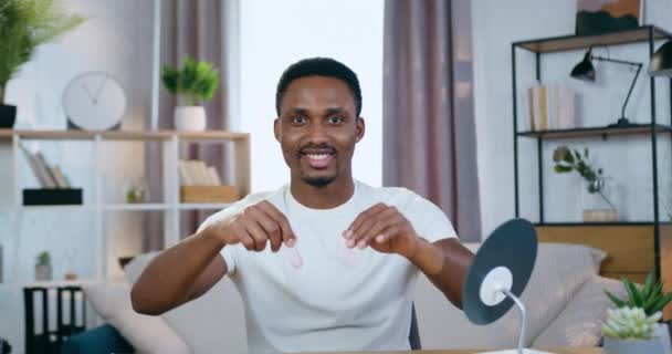 Bonito sorriso despreocupado jovem afro-americano sentado na frente da câmera em casa e mostrando manchas refrescantes de colágeno sob os olhos — Vídeo de Stock