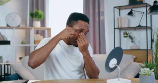 Atraente positivo moderno jovem afro-americano tentando colocar manchas rosa refrescantes sob os olhos olhando para o pequeno espelho em casa, conceito de cuidados com o rosto — Vídeo de Stock