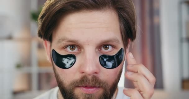 Face care concept where likable smiling caring young bearder touching black hydrogel patches under eyes and looking into camera — Stock Video