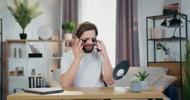 Симпатичный 30-летний носитель с коллагеном освежающими пятнами под глазами, сидящий перед маленьким зеркалом дома и разговаривающий по мобильному телефону — стоковое видео