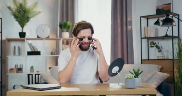 Handsome positive 30-aged bearder with collagen refreshing patches under eyes sitting in front of small mirror at home and talking on mobile — Stock Video