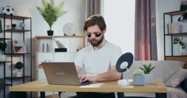 Симпатичный улыбающийся современный носитель 30-летнего возраста с гидрогелевыми повязками, сидящими за столом и работающими на компьютере, крупным планом — стоковое видео