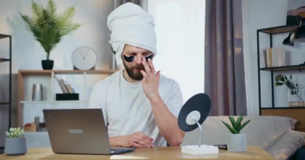Front view of good-looking smiling caring young bearder with hydrogel black patches under eyes which sitting with towel on his head in front of small mirror and looking at his reflection — Stock Video
