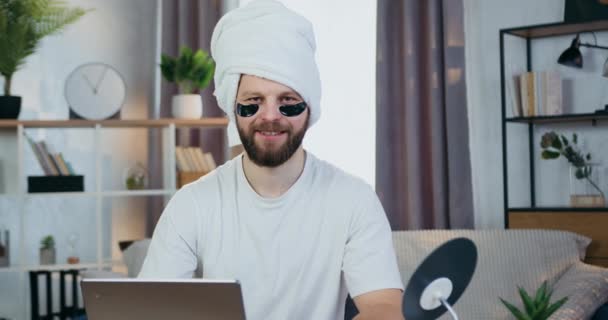 Attractive smiling modern young bearder looking into camera with collagen refreshing eye-patches and towel around his head,beauty concept — Stock Video