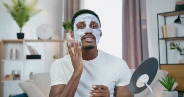 Gesichtspflegekonzept gutaussehende fürsorgliche junge Afroamerikanerin trägt Gesichtsmaske auf, wenn sie zu Hause vor der Kamera sitzt — Stockvideo