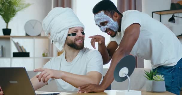 Attractive cheerful young multiracial two male friends with eye-patches and facial mask laughing when revisioning amusing videos on computer at home — Stock Video