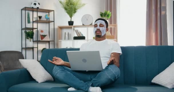 Vista frontal de buen aspecto tranquilo joven de piel negra moderna con máscara facial blanca que se sienta en la pose de loto en el sofá con el ordenador portátil y meditar con los ojos cerrados — Vídeos de Stock