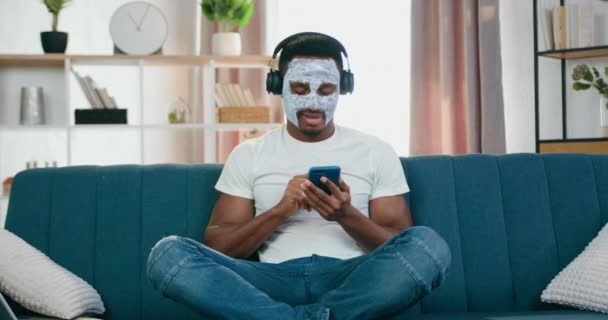 Agradable relajado joven afroamericano moderno con máscara facial sentado en la pose de loto en el sofá y disfrutar de la música favorita en los auriculares — Vídeos de Stock