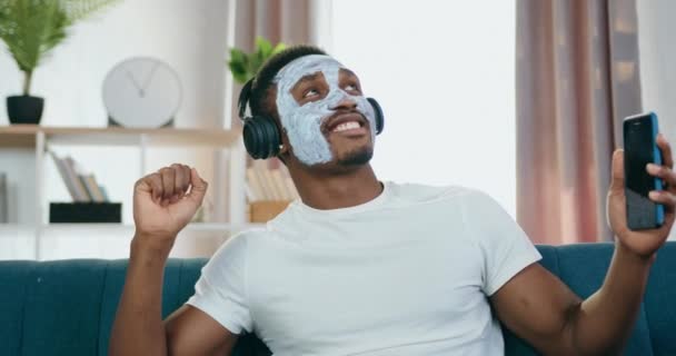 Probable drôle jeune homme à la peau noire moderne dans les écouteurs porte masque hydratant et profiter de la musique de danse à la maison — Video