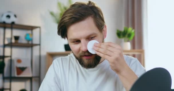 Attraente moderno felice sorridente giovane portatore guardando nello specchio mentre pulisce la sua pelle del viso e la pelle intorno agli occhi con batuffolo di cotone, concetto di procedure facciali — Video Stock