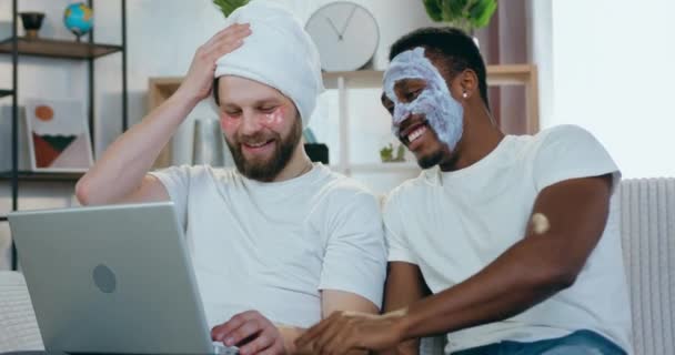 Atractivo sonriente jóvenes hombres modernos multiétnicos viendo videos divertidos en el ordenador portátil, mientras que hizo los procedimientos cosméticos faciales en casa, vista frontal — Vídeos de Stock