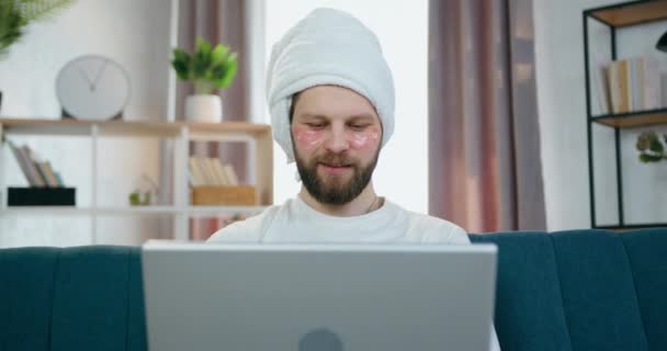 Front view of good-looking positive young bearded guy with pink collagen eye-patches and shower towel on head which sitting on the home couch and working on laptop — Stock Video