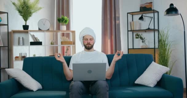 Przystojny, spokojny, młody, opiekuńczy brodzik z przepaskami na oko i białym ręcznikiem wokół głowy, siedzący przed laptopem na kanapie i medytujący zamkniętymi oczami — Wideo stockowe