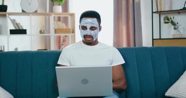 Привлекательный современный заботливый молодой африканский американец с освежающей белой маской для лица сидит на диване дома и использует компьютер, вид спереди — стоковое видео