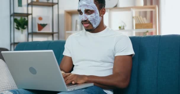 Beau positif jeune homme à la peau noire moderne avec masque hydratant assis devant un ordinateur portable sur le canapé et bavarder avec des amis — Video