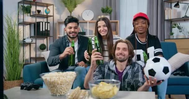 Likable χαρούμενα ανέμελη νέους πολυεθνικούς ανθρώπους clinking μπουκάλια μπύρας κατά τη διάρκεια της αναθεώρησης ποδοσφαιρικό αγώνα στην τηλεόραση στο σπίτι, αναψυχής έννοια — Αρχείο Βίντεο