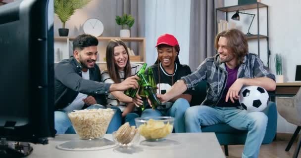 Beautiful two multiracial girls and handsome two diverse guys knocking bottles of beer while sitting on the sofa in front of TV set to watch sports or to play games — стоковое видео