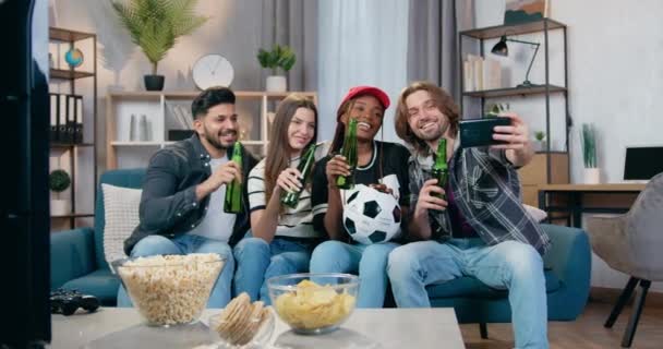 Agradable sonrisa feliz cuatro amigos de raza mixta sentado en el sofá en casa con botellas de cerveza y hacer selfie durante el partido de fútbol revisión en el televisor — Vídeo de stock