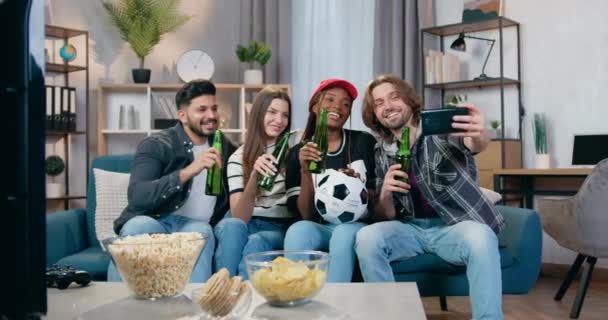 Sympathisch lächelnd glücklich sitzen vier gemischte Rassenfreunde zu Hause mit Bierflaschen auf der Couch und machen Selfie während eines Fußballspiels am Fernseher — Stockvideo