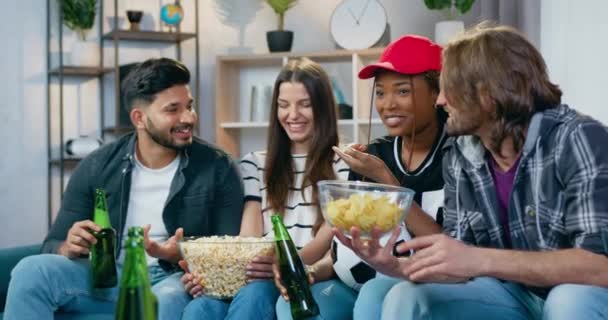 Atrakcyjny pozytywny szczęśliwy 25 różnych przyjaciół siedzi na kanapie w domu i mówi o meczu piłki nożnej oglądają w telewizji, ciesząc się chipsy, popcorn i piwo — Wideo stockowe