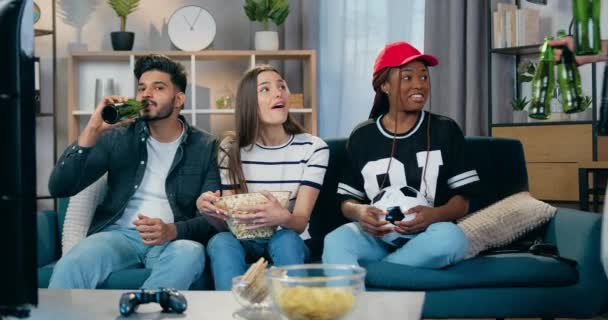 Attractive positive young multiracial three friends watching sports game on TV set when their joint male mate give them bottles of beer and joining to them — Stock Video