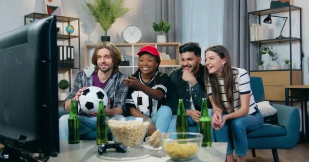 Atractiva sonrisa satisfecha joven mixta gente divirtiéndose juntos mientras ven el juego de deportes en la televisión en casa con aperitivos y cerveza — Vídeos de Stock