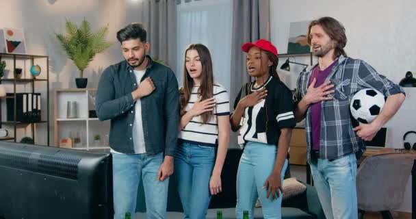 Goed uitziende zelfverzekerde jonge gemengde ras vrienden staan voor TV thuis en zingen patriottische hymne voor het kijken naar wedstrijd, slow motion — Stockvideo