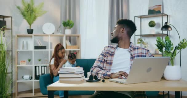 Afrikansk man arbetar på laptop medan frun bryr sig om barn — Stockvideo