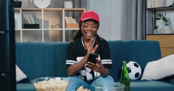 Красивая улыбающаяся счастливая молодая африканская американка в спортивной одежде сидит дома на диване и разговаривает по телефону по видеосвязи — стоковое видео