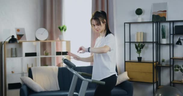 Treadmill σπίτι προπόνηση όπου όμορφος χαρούμενος χαμογελαστός λεπτή νεαρή γυναίκα κάνει τρέξιμο ασκήσεις για το τρέξιμο κομμάτι και ρύθμιση του προγράμματος στο ταμπλό — Αρχείο Βίντεο