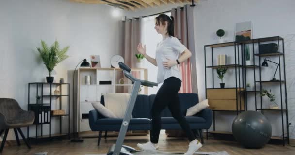 Entrenamiento en casa cardiovascular donde encantadora mujer joven en forma activa trotando en la cinta de correr en casa y comprobar los indicadores en su smartwatch, vista lateral — Vídeos de Stock