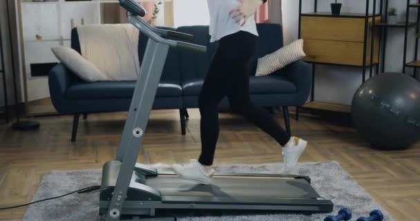 Концепция здорового образа жизни, где довольно активная стройная молодая женщина в спортивной одежде тренируется на беговой дорожке дома и контролирует индикаторы на своих умных часах — стоковое видео