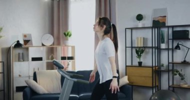 Kardiyo ev antrenmanı. Evde koşu bandına basarken çekici, narin, genç esmer kadın rahatlama egzersizleri yapıyor.