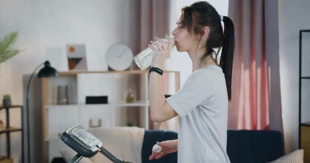 Прекрасная улыбающаяся спортивная молодая женщина с хвостиком пьет воду из бутылки, чтобы пополнить водный баланс в организме во время тренировок на беговой дорожке дома — стоковое видео