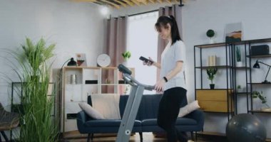 Evde koşu bandında yürüyen ve akıllı telefonunu Freinds 'le sohbet etmek için kullanan çekici, güler yüzlü, narin, aktif genç bir kadının yan görüntüsü.