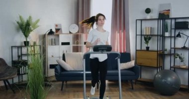 Kardiyo ev egzersizleri konsepti. Güzel görünümlü, kendine güveni olan, 25 yaşında, rahat bir dairede koşu bandında koşan bir kadın.