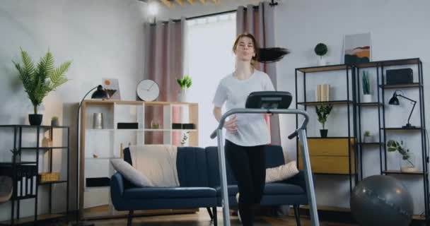 Mujer deportiva delgada bastante positiva en ropa de entrenamiento que corre rápido en la cinta de correr durante el entrenamiento en casa interior, vista frontal, cámara lenta — Vídeos de Stock