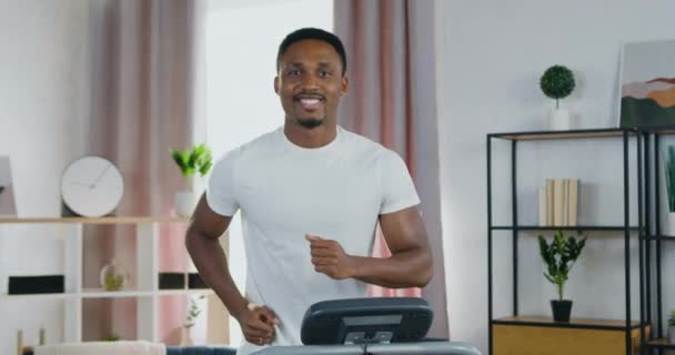 Привлекательный улыбающийся спортивный молодой африканский американец смотрит в камеру во время тренировки на беговой дорожке дома, концепция здорового образа жизни — стоковое видео