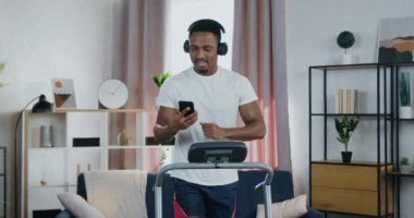 Evde egzersiz konsepti. Yakışıklı, gülümseyen, aktif, genç, siyah tenli bir adamın evde koşu antrenmanında olması ve favori şarkıların kulaklıkla dinlenmesi.