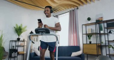 Spor giyimli, yakışıklı, gülümseyen genç, aktif, siyah tenli bir adam. Evdeki koşu bandında antrenmanlar ve kulaklıkla modern müzik dinlemek.