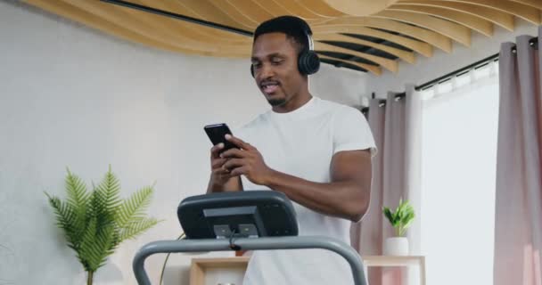 Likable positive active young black-skinned guy running on treadmill during home training and enjoying favourite melodies in headphones from phone playlist — Stock Video